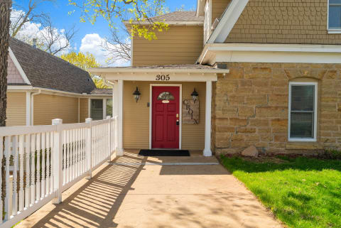 Charming two-story home at 305 featuring a welcoming red front door, stone and wood siding, and a white picket fence on a sunny spring day.