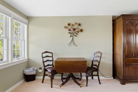A small dining area with a drop-leaf table, two wooden chairs, and floral wall art, illuminated by natural light from the windows.