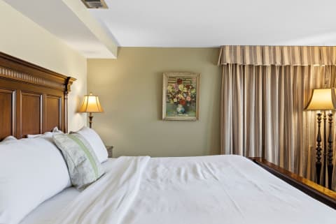 A serene bedroom with a plush bed, decorative pillow, lamp, floral painting, and flowing curtains.