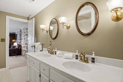 A luxurious ensuite bathroom with dual sinks, ornate mirrors, and soft lighting opening to a cozy sitting area.