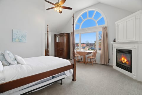 A luxurious suite with a four-poster bed, cozy fireplace, and large arched window overlooking a vibrant street.