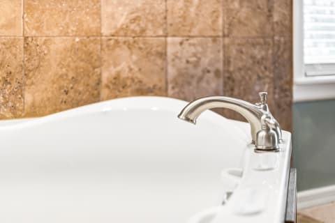 A white soaking tub with a sleek silver faucet against warm travertine tiles.