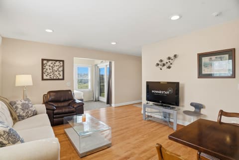 A cozy living room with a floral couch, brown recliner, and a TV; bright with natural light.