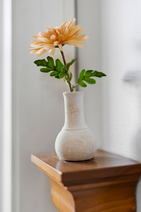 A small peach flower in a textured ceramic vase on a wooden shelf.