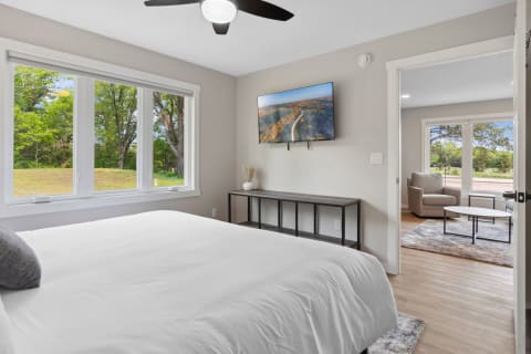 Modern bedroom featuring a view of trees, a white bed, and a small living area in the background.