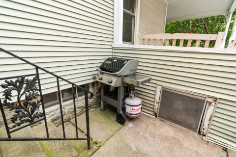 Gas grill next to a green house with a black iron handrail.