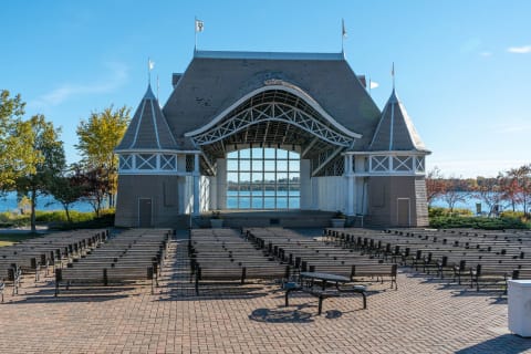 Outdoor amphitheater with a curved roof and wooden benches, surrounded by autumn foliage and a lake.