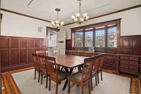 A warm and inviting dining room with a large wooden table, surrounded by chairs and illuminated by chandeliers, with beautiful stained glass windows.