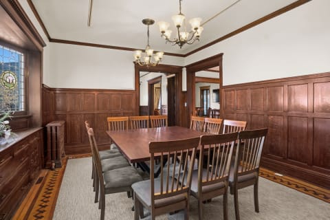 A sophisticated dining room with wooden paneling, a long table with chairs, and elegant chandeliers, illuminated by a stained glass window.