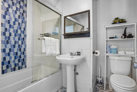 A stylish bathroom with hexagonal blue tiles, glass-enclosed tub, pedestal sink, and organized shelves.