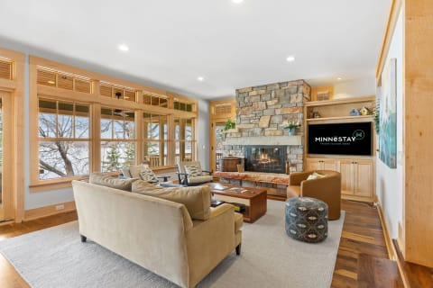 Interior view of a living room with a stone fireplace and large windows overlooking trees.