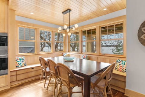 A bright dining area with a wooden table, rattan chairs, and colorful cushions, surrounded by large windows.