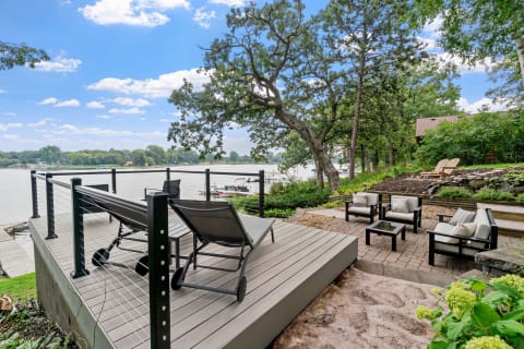 A lakeside deck with loungers and seating area surrounded by greenery and a calm lake.