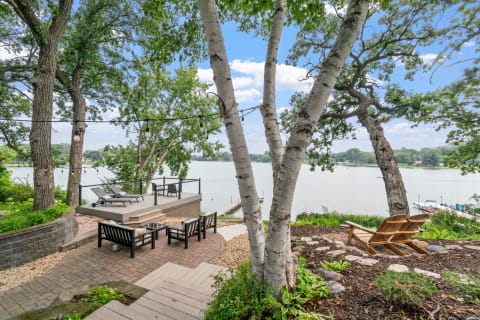 Lakeside patio view with seating, lounge chairs, and string lights among trees.