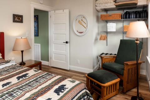 A cozy bedroom with a patterned quilt, a lamp on a nightstand, and a reading nook with a green chair.
