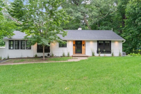 A charming white house with dark trim and a wooden front door, set in a grassy yard amidst trees.