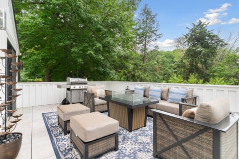 An elegant outdoor patio with a glass table, cushioned seating, and a grill, set amidst greenery.
