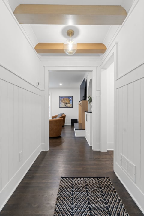 A modern hallway with white walls, wooden beams, and a cozy living area visible in the background.