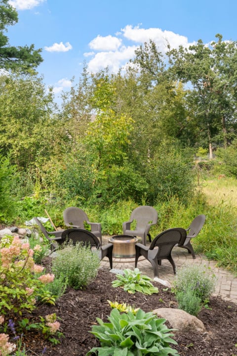 A circular arrangement of dark chairs around a fire pit in a lush garden.