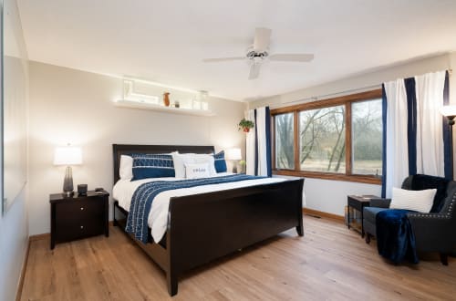 Unwind in your serene bedroom where navy accents meet crisp whites, while natural light streams through large windows overlooking peaceful wooded views.