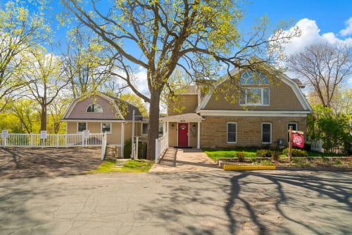 Historic stone farmhouse with modern amenities, nestled among mature trees with ample parking and a welcoming red entrance door.